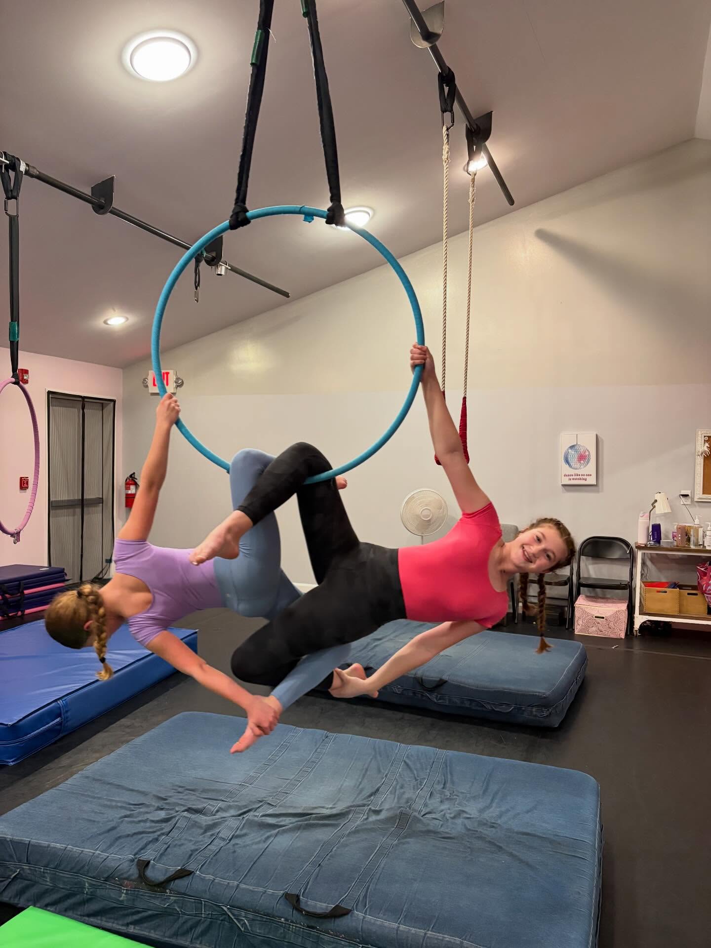 Two students practicing together on a turquoise aerial hoop above a mat
