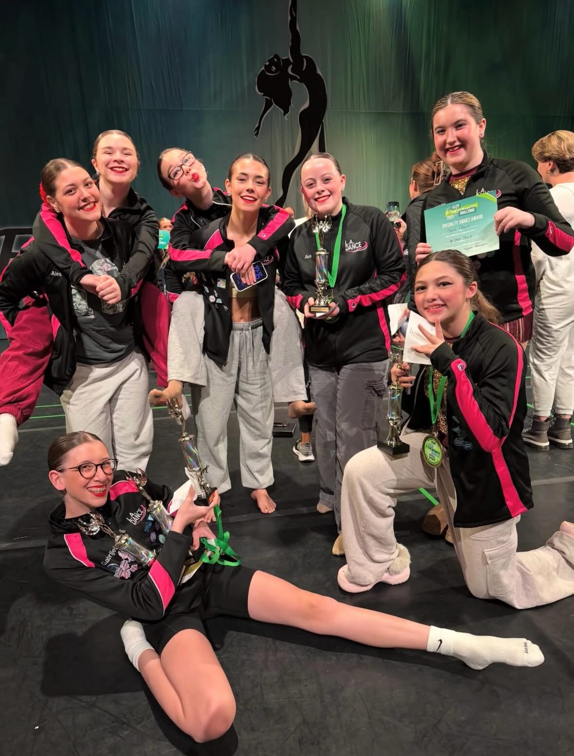 A group of dancers in black jackets and gray pants smiling together backstage